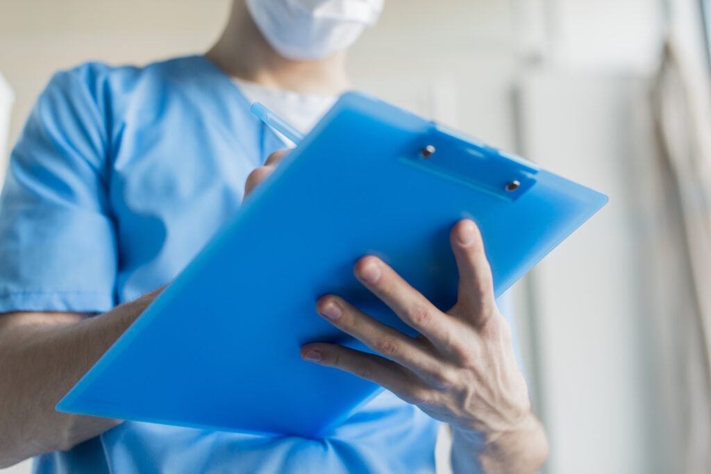 A imagem mostra o tronco de um homem que veste uma roupa azul, que é um uniforme usado por profissionais de Enfermagem. Nas mãos dele há uma prancheta azul, na qual ele faz anotações. 