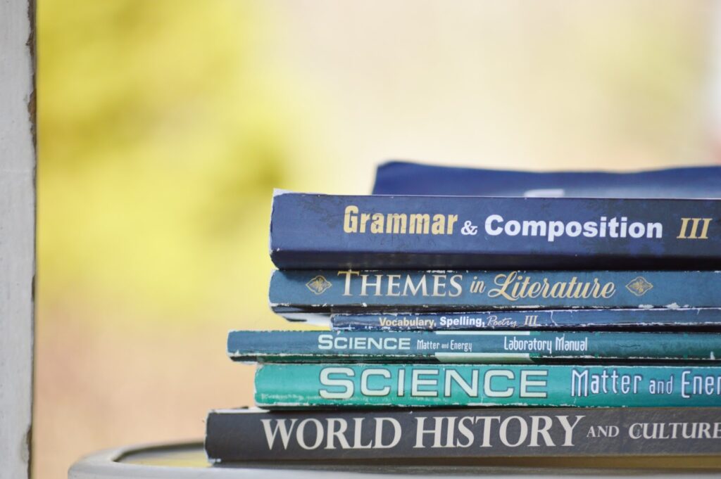 A imagem mostra seis livros empilhados. Todos eles possuem título em inglês e possuem assuntos acadêmicos. De baixo para cima, há um livro da história do mundo e cultura, dois livros de ciência, um de vocabulário e fala, outro sobre temas na literatura e um último sobre gramática e escrita. 