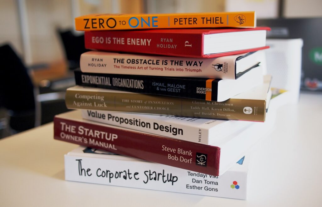 La imagen muestra ocho libros apilados sobre una mesa blanca. Es posible ver sus lomos, en los que están escritos sus títulos en inglés: “Zero to one”; “Ego is the enemy”; “The obstacle is the way ”; “Exponential organizations”; “Competing against luck”; “Value Proposition Design”; “The Startup owner’s manual”; “The corporate startup”.