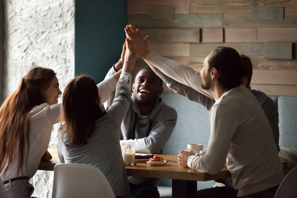 A imagem mostra um grupo de pessoas reunido em torno de uma mesa de café. Da esquerda para a direita, há uma mulher branca ruiva, ao lado dela há outra mulher branca, de cabelo castanho liso, um homem branco de cabelo castanho, um homem branco que não aparece tanto na imagem e um homem negro sorrindo. Todos estão com um dos braços levantados e as mãos deles se encontram no centro da mesa, como em um sinal de união.