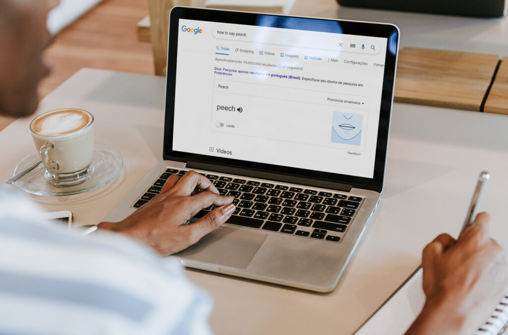 A imagem mostra em destaque um laptop prata sobre uma mesa branca. Na tela dele, é possível ver a tela de busca do Google, com a palavra peach destacada. Também é possível ver a mão esquerda de uma pessoa negra no teclado do notebook. A mão direita dela está segurando uma caneta sobre um caderno de anotações. 