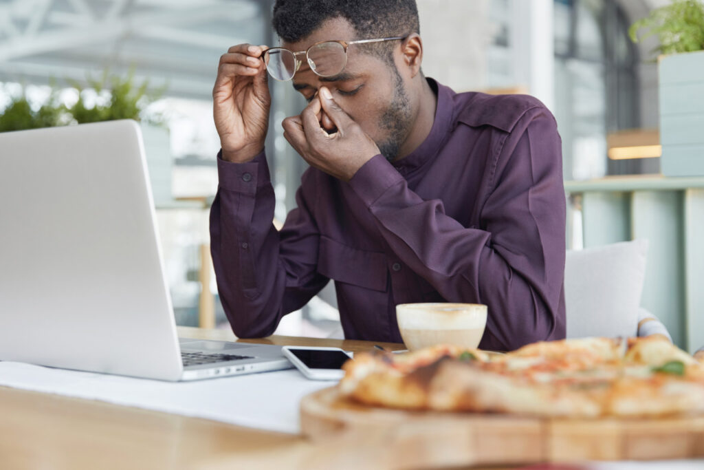 A imagem mostra um homem negro, vestindo camisa social vinho, sentado em frente a uma mesa de madeira. Com a mão direita ele segura os seus óculos acima da testa. A mão esquerda dele está pressionando a área entre os olhos e o nariz, como se estivesse preocupado. 