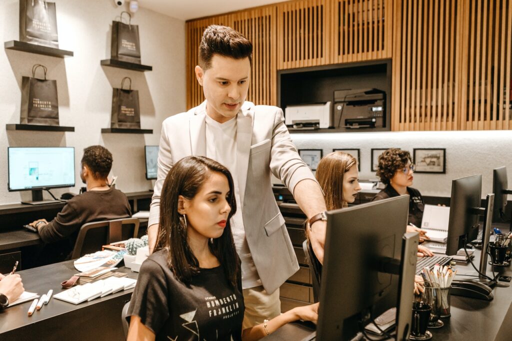 La imagen muestra un grupo de personas en una sala de trabajo con escritorios y computadores. En primer plano, es posible ver a una mujer blanca con el pelo castaño liso a la altura de los hombros sentada frente a un monitor. A su izquierda, hay un hombre blanco de pie. Él señala la pantalla del computador y parece ayudar a la mujer en alguna tarea del trabajo.