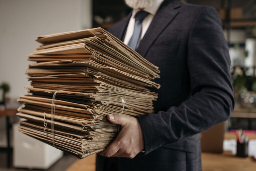  A imagem mostra um homem de terno preto e gravata azul segurando uma pilha de documentos na mão.