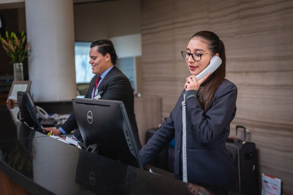 A imagem mostra duas pessoas, uma mulher e um homem, na recepção de um hotel. Eles estão no lado direito da imagem e é possível vê-los na parte de dentro do balcão e vestem terno, que aparenta ser o uniforme dos trabalhadores do local. A mulher é branca, tem cabelo castanho liso, que aparece preso. Ela está em frente a um monitor e com a mão esquerda segura um telefone branco na altura da orelha. Ao lado dela, em menor destaque, está o homem branco, de cabelo preto liso. Ele está em frente a um monitor de computador e sorri. 