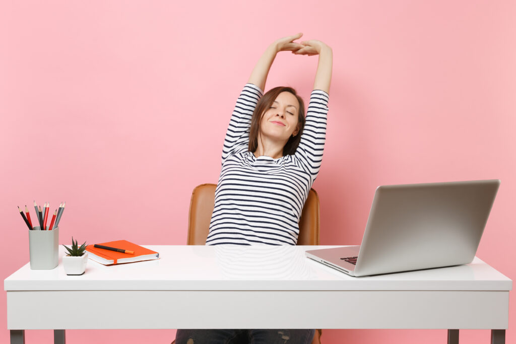 A imagem mostra uma mulher branca de cabelo castanho liso. Ela está sentada em frente a uma mesa branca, onde é possível ver um laptop prata, um porta-lápis, um vasinho de flor e um caderno de anotações. A mulher está com os braços erguidos acima da cabeça, como se estivesse se alongando.