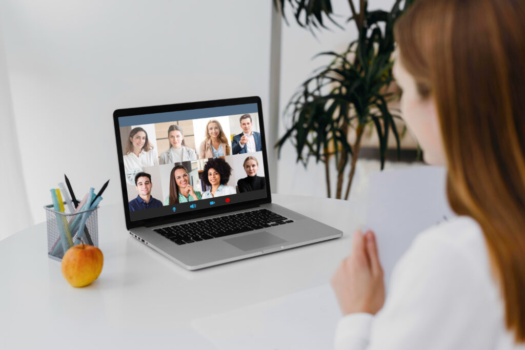 A imagem mostra uma mulher branca, de cabelo castanho-claro liso, em frente a um notebook prata. Na tela dele, é possível ver a imagem de outras pessoas em uma videoconferência.