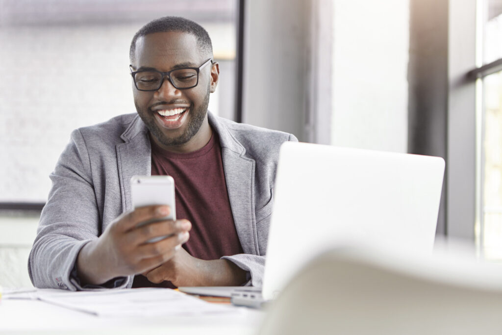 A imagem mostra um homem negro sentado em frente a uma mesa de trabalho. Ele usa óculos quadrados pretos, uma camiseta vermelha e um blazer cinza. Na mão esquerda, ele segura um celular prata. Seu olhar está voltado para a tela do aparelho e ele está sorrindo. Em menor destaque na imagem é possível ver um laptop e alguns papéis em cima da mesa. 