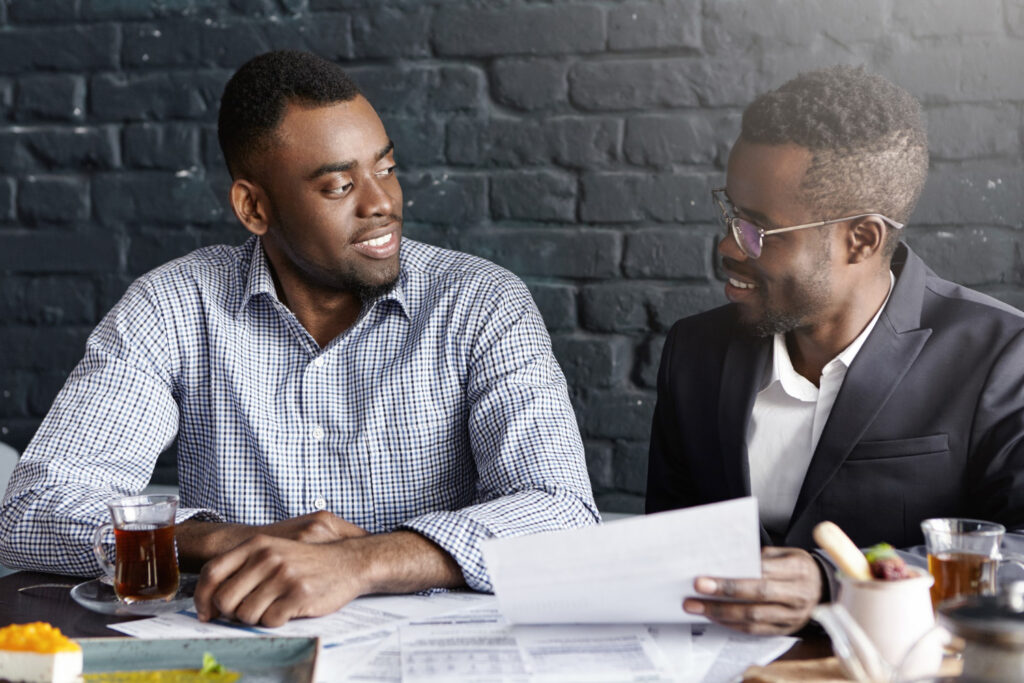 Dois homens negros estão em frente a uma mesa, na qual há alguns papeis e xícaras. Eles se olham e sorriem. 