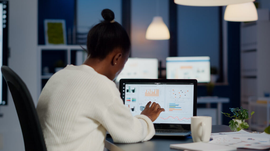 A imagem mostra uma mulher negra, de cabelo preto que está em um coque. Ela está de costas para a foto e aparece sentada em frente a uma mesa de escritório. A mulher mexe em um laptop preto e, na tela dele, é possível ver alguns gráficos coloridos, como se fosse um relatório de resultado. 
