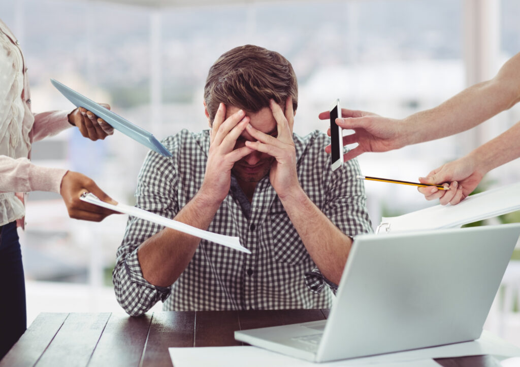 Um homem branco em frente a uma mesa está com as mãos no rosto. Ao redor dele aparecem mais quatro mãos, cada uma segura uma coisa: papéis, lápis, telefone. Ele parece estar sobrecarregado de trabalho. 