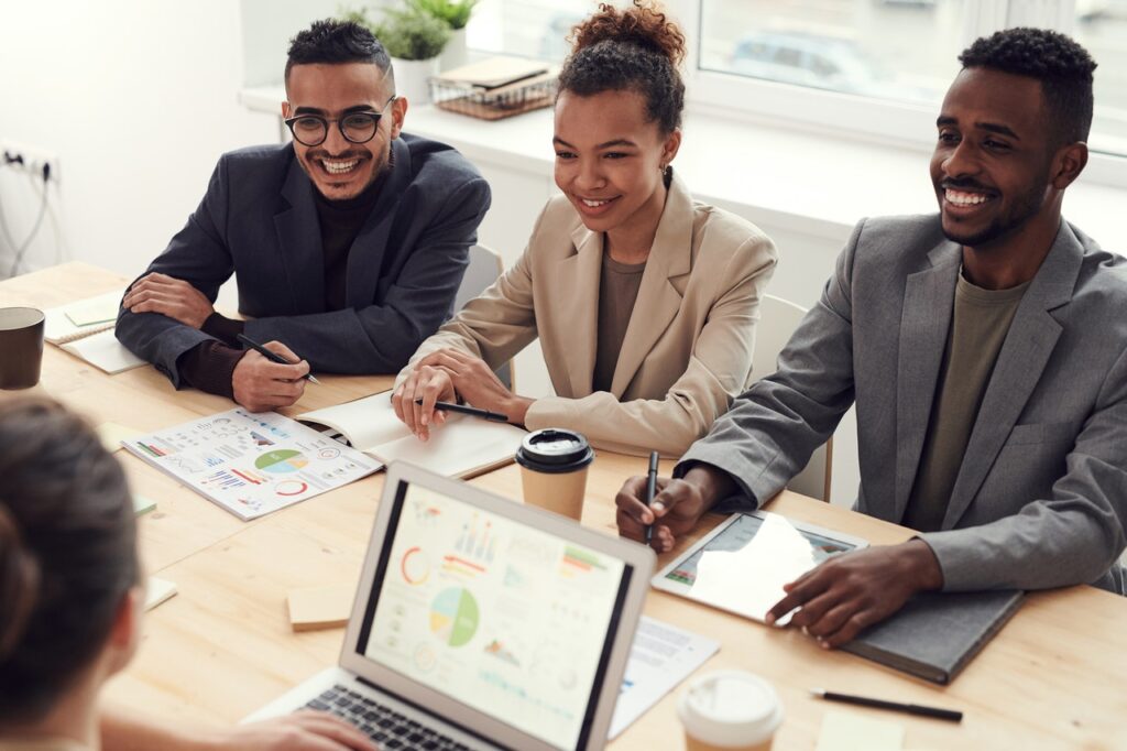 Um grupo de pessoas está em torno de uma mesa de madeira. Da esquerda para a direita há um homem branco, uma mulher negra e um homem negro. Também há uma mulher branca que aparece de costas e desfocada na imagem. Eles sorriem e parecem felizes com o emprego, o que é proporcionado pelo upskillig e reskilling.