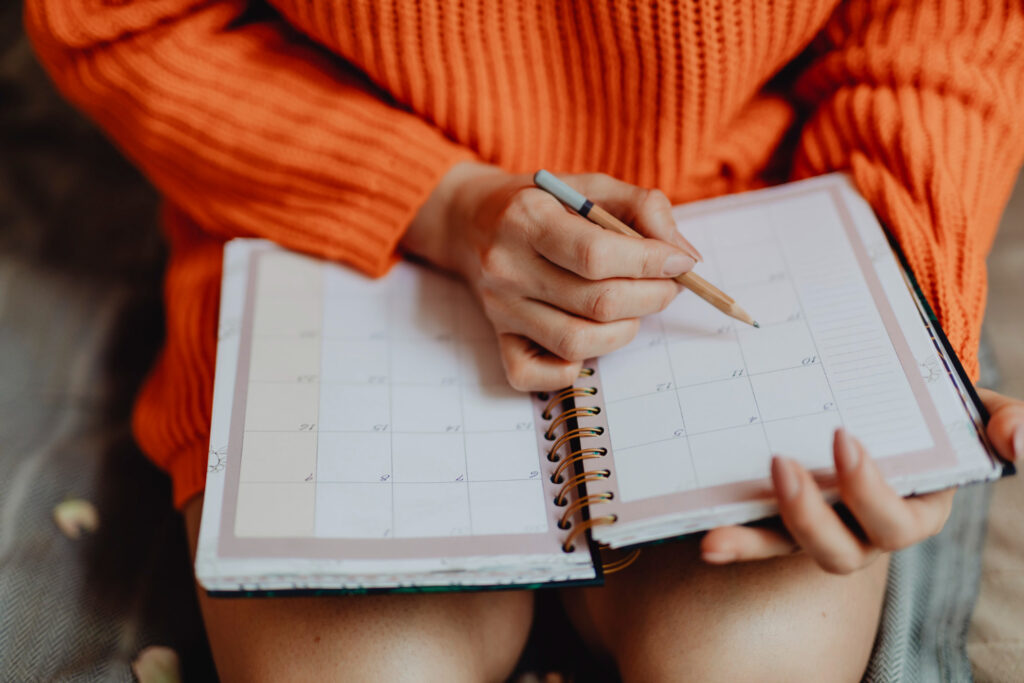 Uma pessoa segura uma agenda aberta e um lápis, com o qual ela anota os compromissos no papel. 