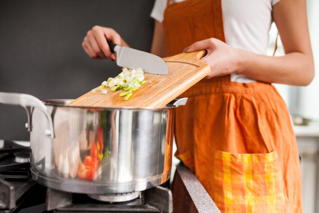 La imagen muestra el torso de una mujer frente a una estufa. Ella lleva un delantal naranja encima de una camiseta blanca. Con la mano izquierda, sostiene una tabla de madera, en la cual se pueden ver algunos ingredientes. En su mano derecha, hay un cuchillo con el que empuja los ingredientes dentro de una olla de acero inoxidable.