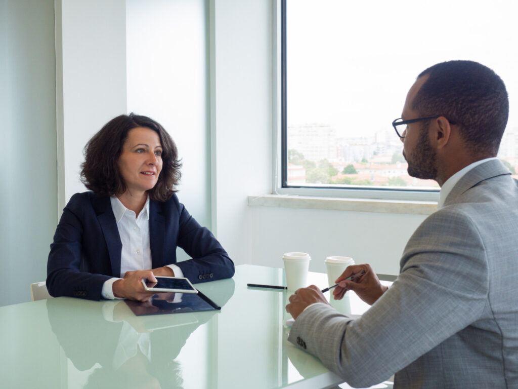 La imagen muestra a un hombre y una mujer sentados frente a una mesa de reuniones blanca. La mujer está en el lado izquierdo de la foto, es blanca y tiene el pelo liso y castaño hasta los hombros. El hombre está frente a ella, es negro y lleva gafas cuadradas negras.