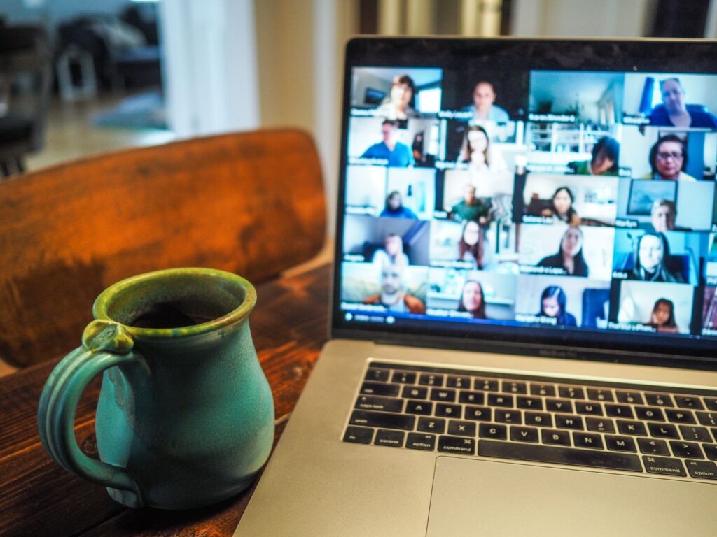 La imagen muestra una laptop plateada  en la que se desarrolla una reunión en inglés, donde es posible ver al menos a 20 perfiles de los participantes. Al lado izquierdo, hay una taza azul verdosa. La laptop y la taza están sobre una mesa de madera.
