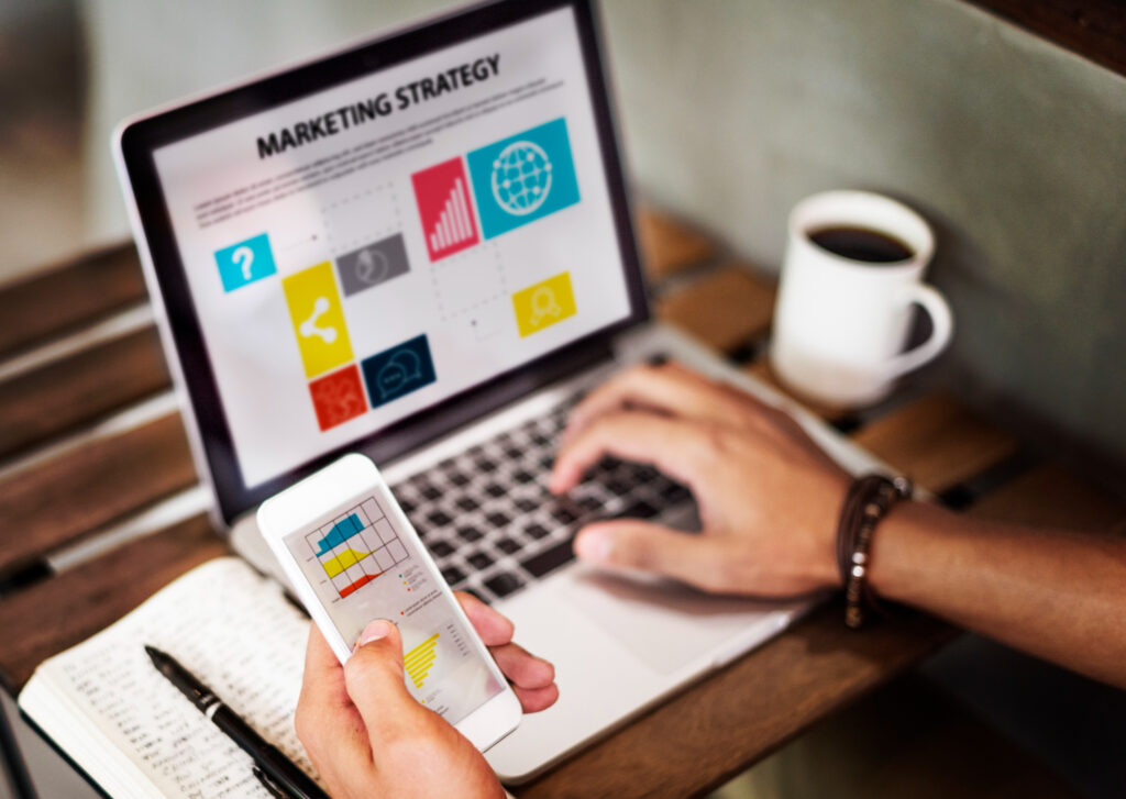 La imagen muestra, en primer plano, la mano izquierda de una persona sosteniendo un celular blanco, en el que se ven unos gráficos de colores. En la parte derecha de la imagen, es posible ver una laptop plateada con la frase “Marketing strategy” en negrilla. La mano derecha de la persona está apoyada sobre el teclado de la computadora.