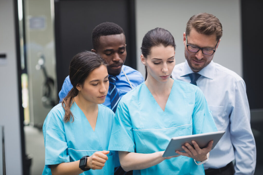 La imagen muestra a un grupo de cuatro personas que trabajan en un hospital mirando una tablet gris; las dos mujeres están en la parte de adelante y los dos hombres están detrás de ellas. De izquierda a derecha, hay una mujer blanca de cabello castaño, liso y recogido en una cola de caballo. Lleva un uniforme de enfermera de color azul. Detrás de ella, hay un hombre negro de cabello crespo que lleva ropa formal. Frente a ella, hay otra mujer blanca de cabello negro, liso y recogido en una cola de caballo. También usa el uniforme de enfermera y sostiene la tablet gris en sus manos. Finalmente, hay un hombre blanco de cabello rubio, liso y gafas negras cuadradas. Lleva camisa formal y corbata.