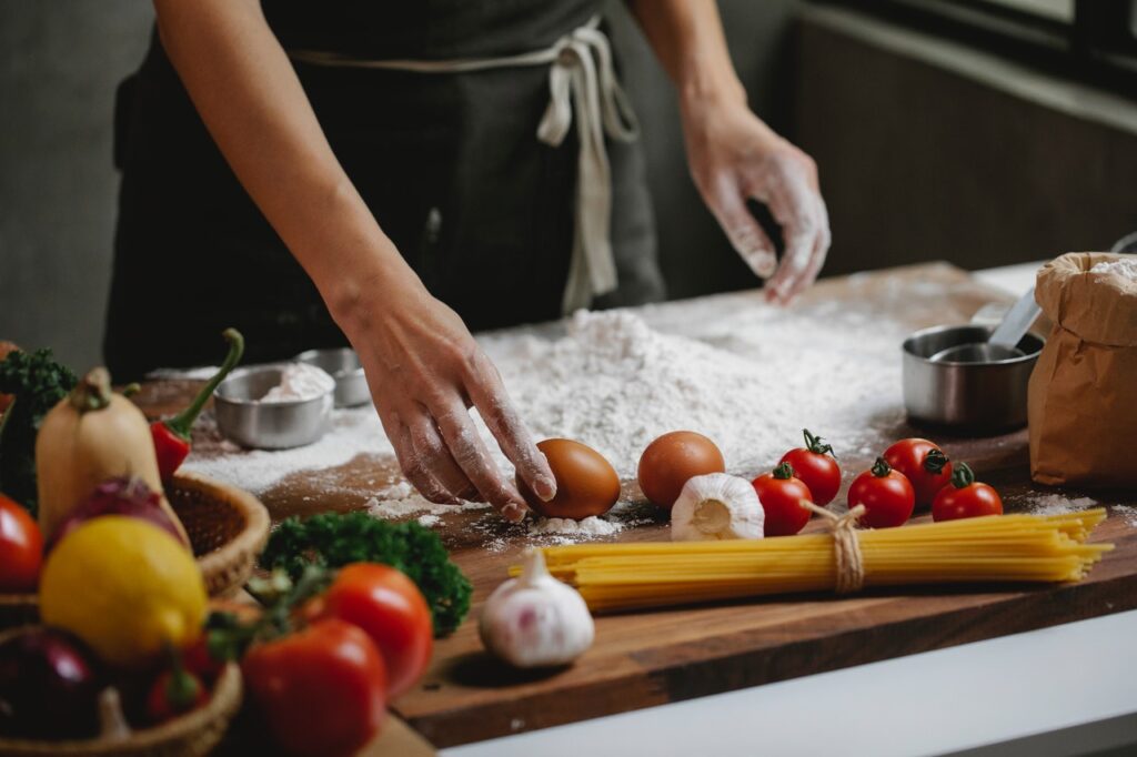 La imagen muestra el tronco de una persona frente a un mesón de cocina con algunos ingredientes. Sobre él, es posible ver un poco de harina de trigo, la mano derecha de la persona está tocando un huevo marrón que está junto a ingredientes como tomates, ajo, pimienta y un poco de espagueti, aún sin cocinar.