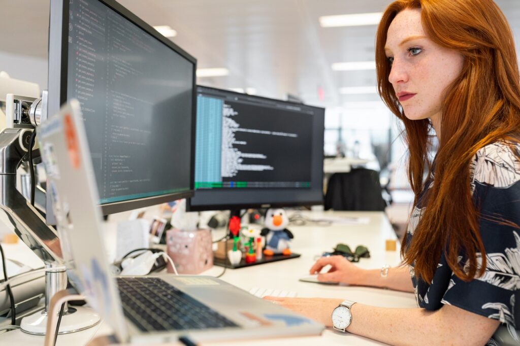 La imagen muestra a una mujer blanca, con el cabello rojo a la altura del pecho, sentada frente a un escritorio. Se le ve de perfil y está ubicada al lado derecho de la imagen. Sobre el escritorio, se ven dos monitores con códigos de programación, una de las profesiones del futuro, y la mirada de la mujer está dirigida hacia ellos. También hay una laptop plateada y algunos adornos encima del escritorio.