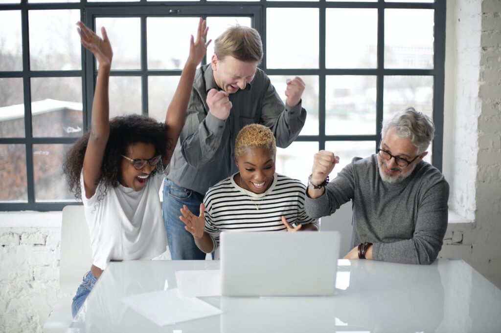 Um grupo de quatro pessoas está reunido atrás de uma mesa branca, onde há um laptop prata. Da esquerda para a direita há uma mulher negra de cabelo cacheado preto, um homem branco de cabelo castanho-claro liso, uma mulher branca de cabelo loiro curto e um homem branco de cabelos brancos. Eles estão felizes e parecem comemorar algo. 