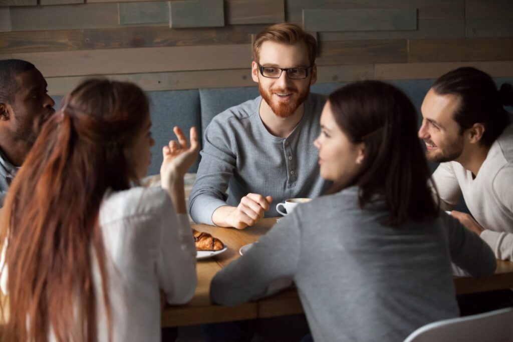 La imagen muestra a un grupo de personas reunidas en torno a una mesa de madera en la que se puede ver un plato con un croissant y un café. De izquierda a derecha, hay un hombre negro, junto a él un hombre blanco pelirrojo, otro hombre blanco con cabello castaño oscuro recogido en un moño. Junto a él, de espaldas a la imagen, hay una mujer blanca con el cabello liso y castaño oscuro, a la altura de los hombros, y otra mujer blanca pelirroja.