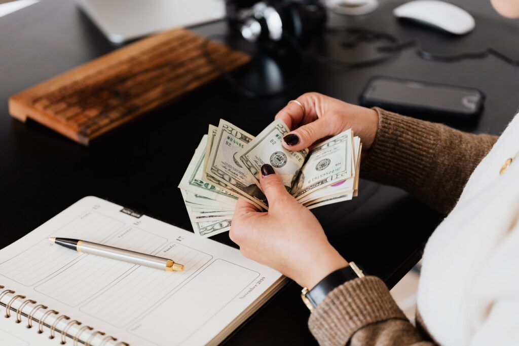 En la esquina izquierda de la imagen, se muestran las manos de una mujer blanca que sostiene unos billetes, como si los estuviera contando. Frente a ella hay una mesa negra con un cuaderno y un bolígrafo.