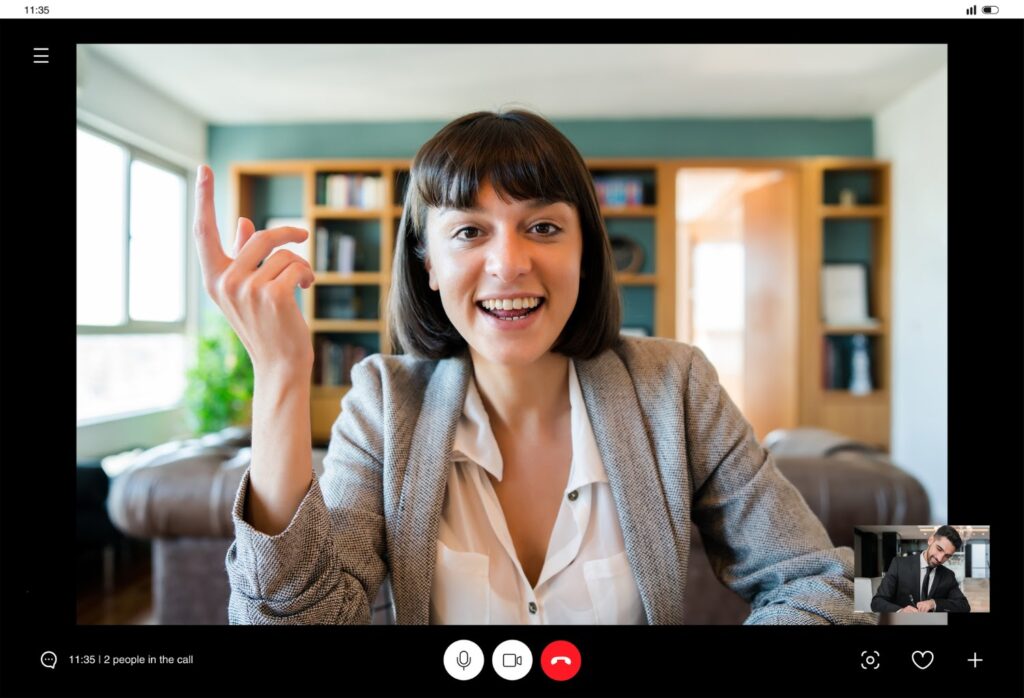 La imagen muestra una captura de pantalla de una videollamada. En ella se puede ver a una mujer blanca con el pelo castaño oscuro a la altura de los hombros y con flequillo. Está sonriendo y su mano derecha está apuntando hacia arriba. En la parte derecha de la imagen, en un recuadro más pequeño, se ve a un hombre blanco con traje, de pelo corto y negro.