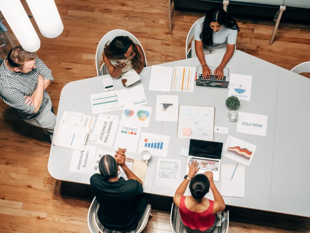 Um grupo de cinco pessoas está em volta de uma mesa de escritório cinza e é visto de cima. Na mesa há alguns papéis, como se o grupo estivesse em uma reunião de ideias. 