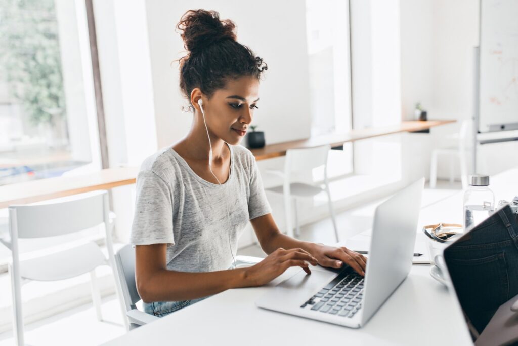 Uma mulher negra, vestindo camiseta cinza, está sentada em uma cadeira, mexendo em um laptop branco, que está apoiado sobre uma mesa. Ela usa fones de ouvido.