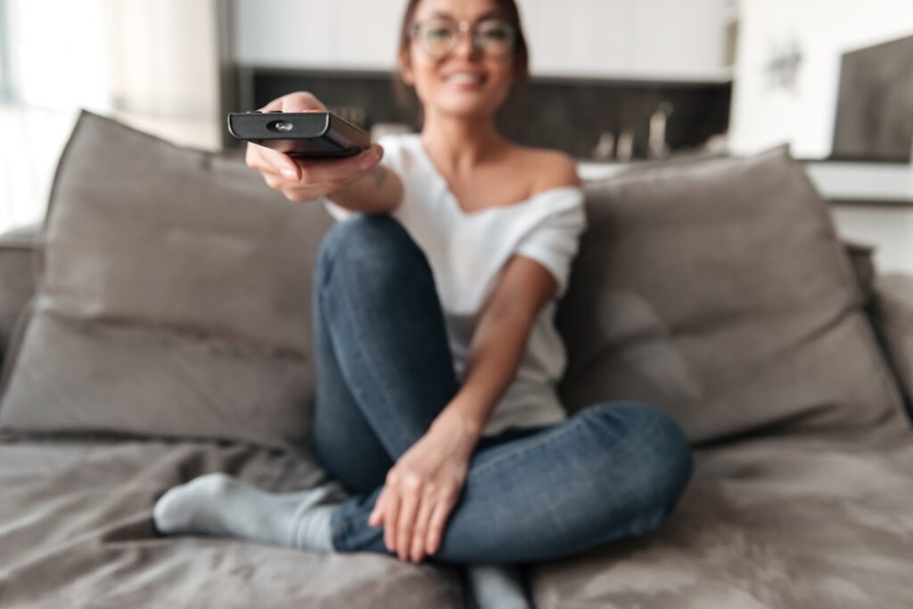 Mulher branca, usando calças jeans e camiseta branca, sentada em um sofá. Com a mão esquerda, ela segura um controle remoto, representando que os programas de televisão em inglês são um caminho de como melhorar o listening.