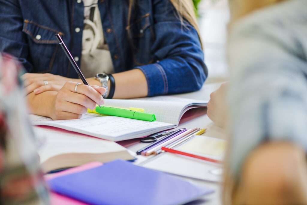 Os braços de uma mulher branca aparecem sobre um caderno. Com uma das mãos, ela segura um lápis preto.