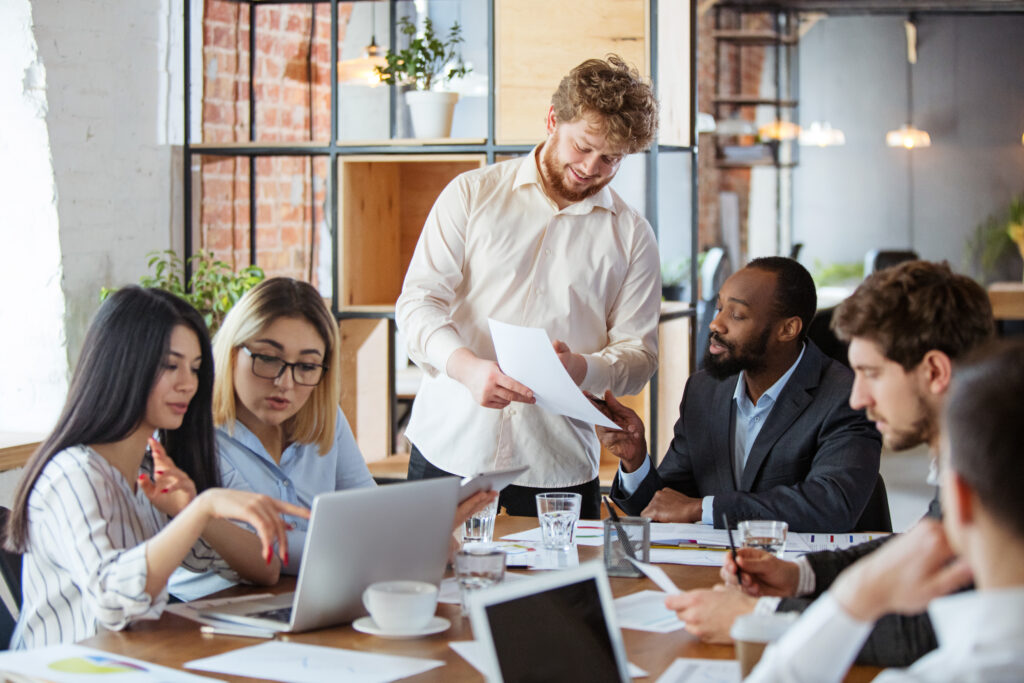 Grupos de pessoas sentadas em uma mesa com várias folhas e laptops conversando sobre trabalho.